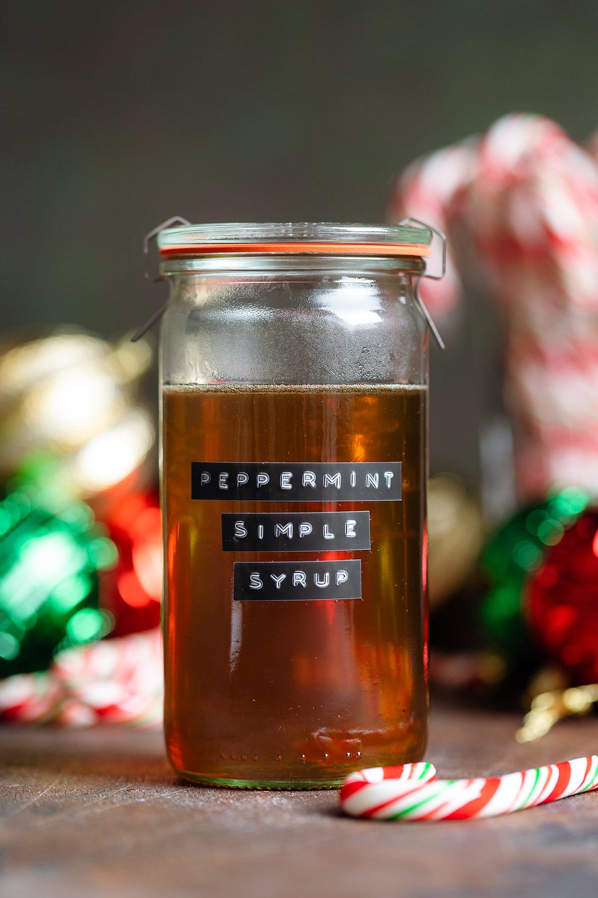 Golden syrup in a glass jar with a glass lid and a black label that says peppermint syrup on the side with Christmas ornaments and candy canes in the background.
