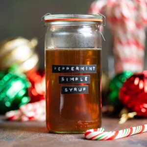 Golden syrup in a glass jar with a glass lid and a black label that says peppermint syrup on the side with Christmas ornaments and candy canes in the background.