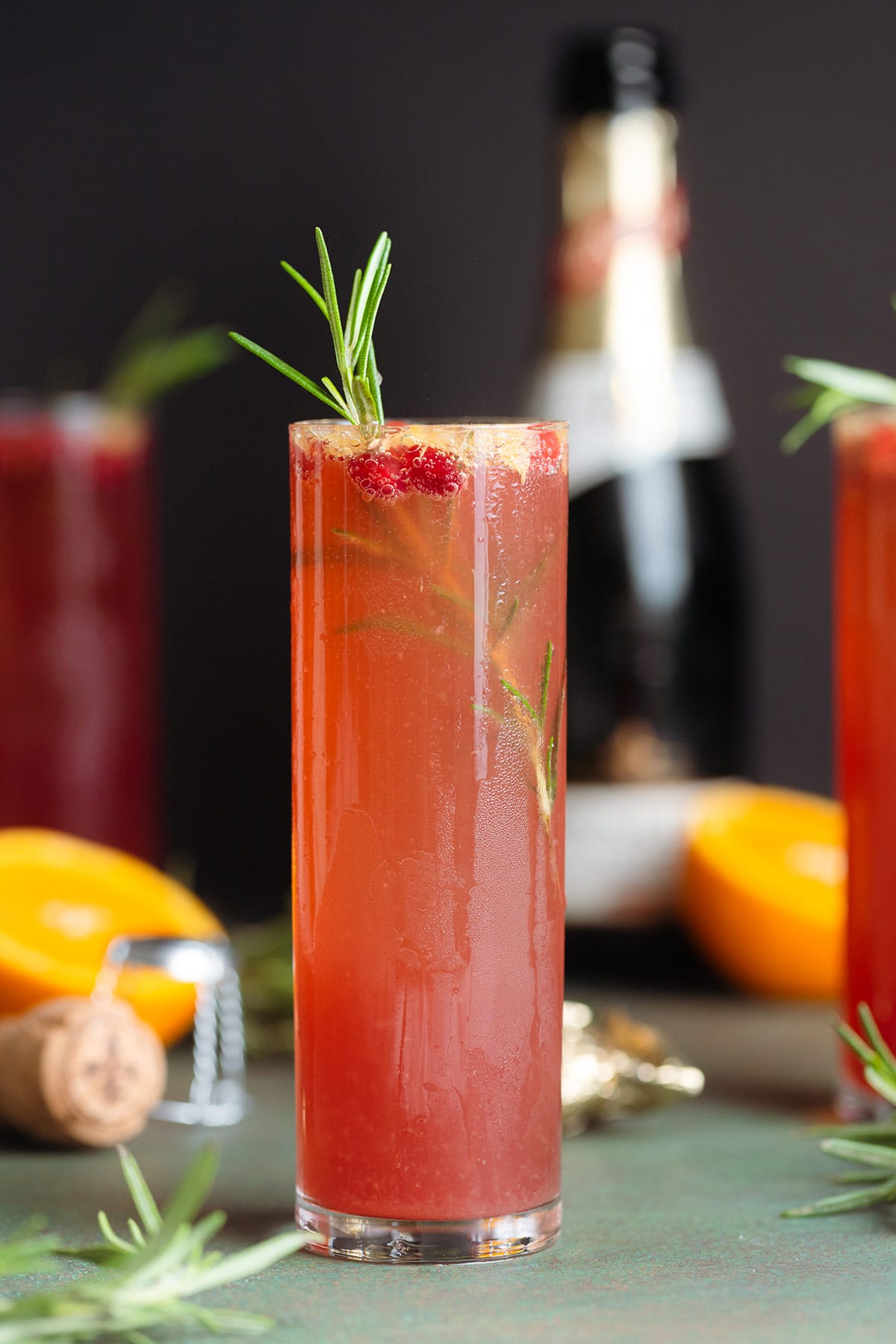 A tall champagne glass with pomegranate mimosa garnished with fresh rosemary and pomegranate arils. with more drinks and fresh orange in the background.