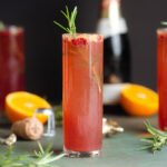 A tall champagne glass with pomegranate mimosa garnished with fresh rosemary and pomegranate arils. with more drinks and fresh orange in the background.