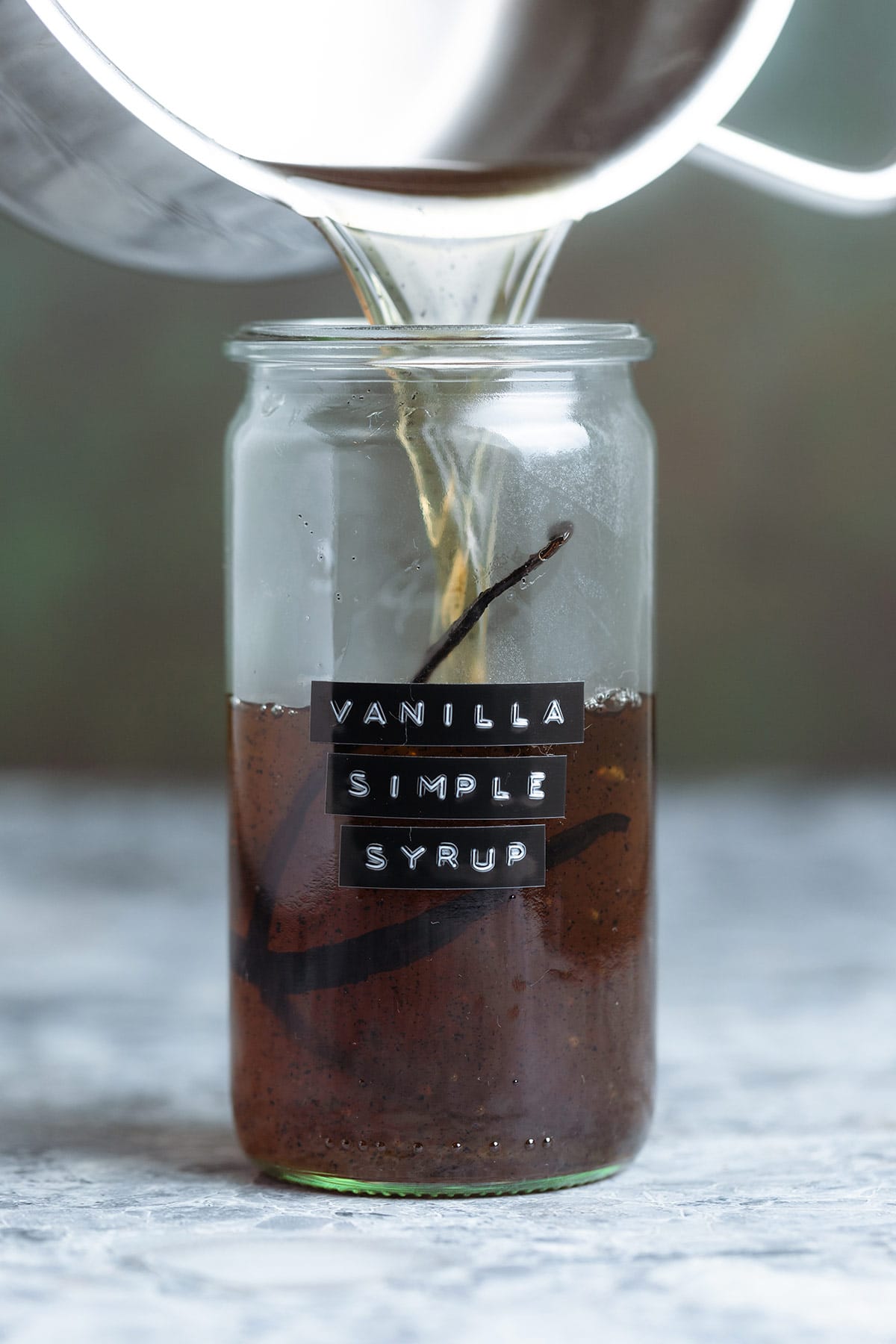 Vanilla syrup being poured into a glass jar with vanilla simple syrup label on it and a whole vanilla bean inside.