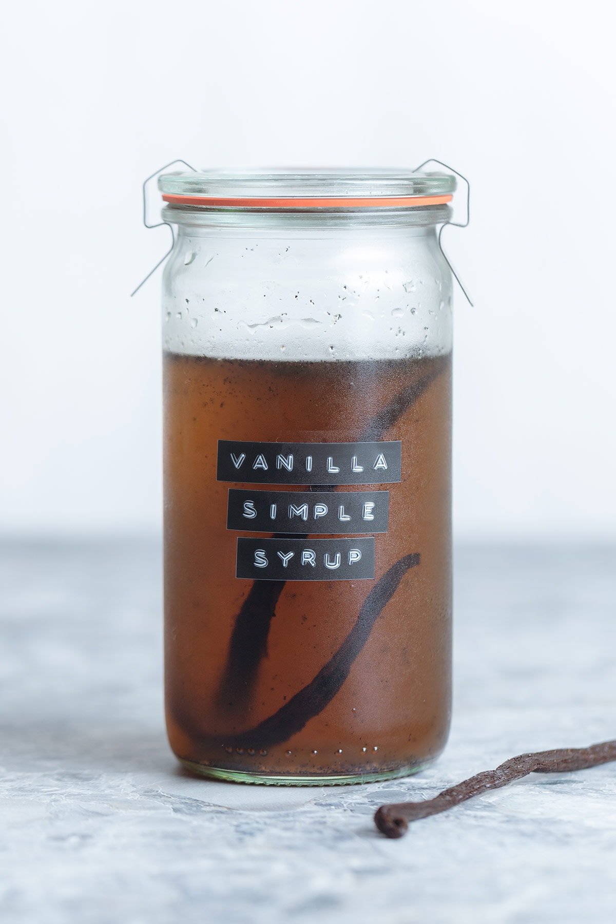A glass jar of vanilla simple syrup with a whole vanilla bean inside the jar on a grey background, the jar has a label on it that says vanilla simple syrup.