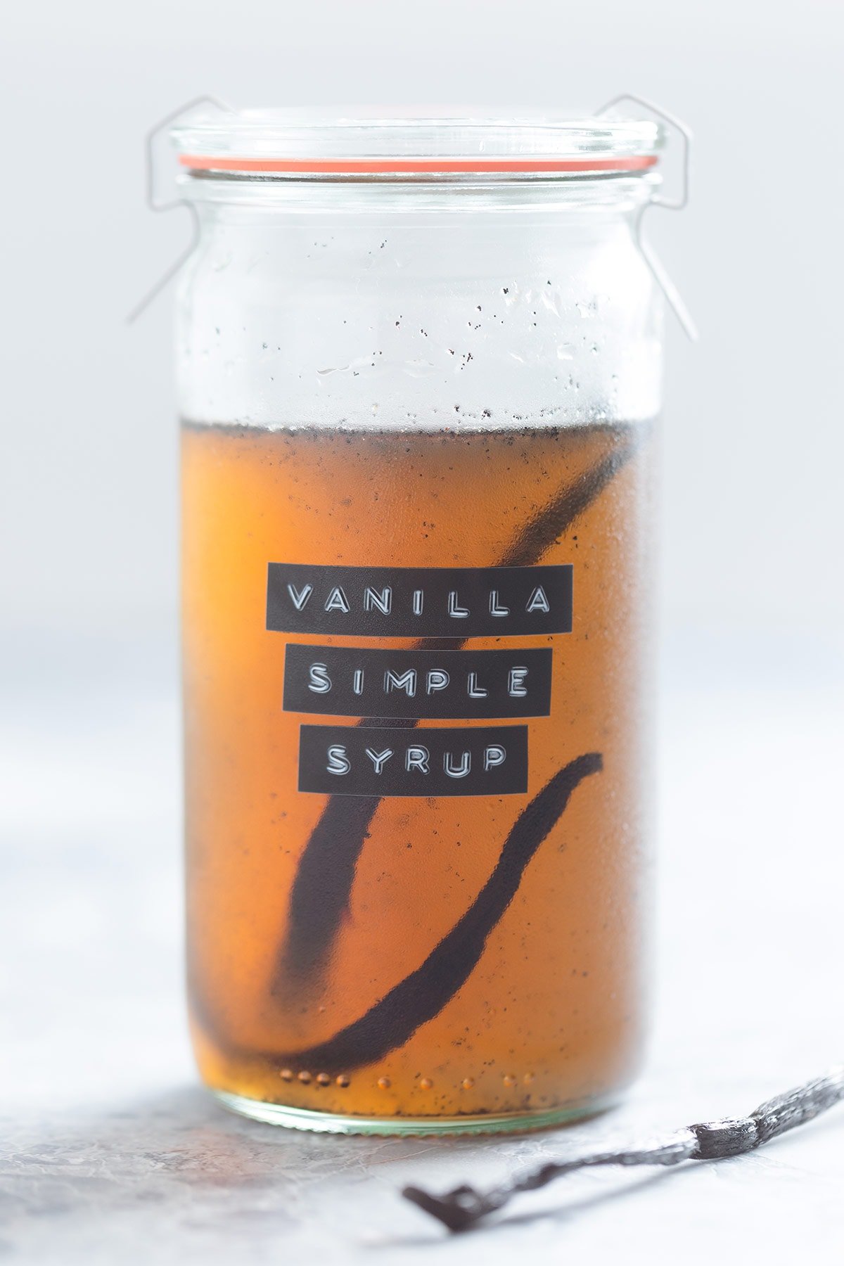 A glass jar of vanilla simple syrup with a whole vanilla bean inside the jar on a grey background, the jar has a label on it that says vanilla simple syrup.
