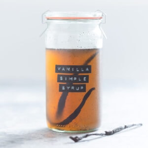 A glass jar of vanilla simple syrup with a whole vanilla bean inside the jar on a grey background.