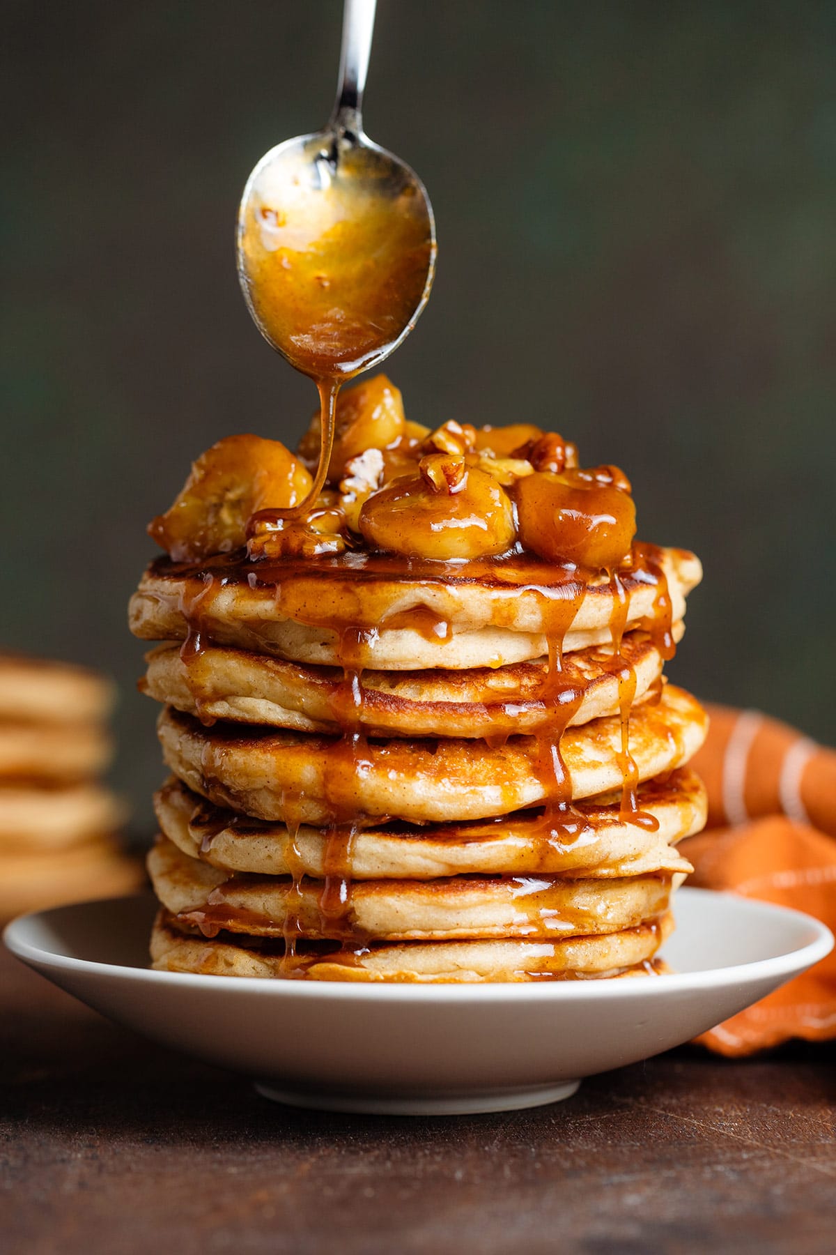 A stack of pancakes with caramelized bananas and pecans with a spoon above drizzling the pancakes with more caramel sauce.