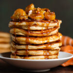 A stack of pancakes with caramelized bananas and pecans with caramel sauce dripping down the sides.