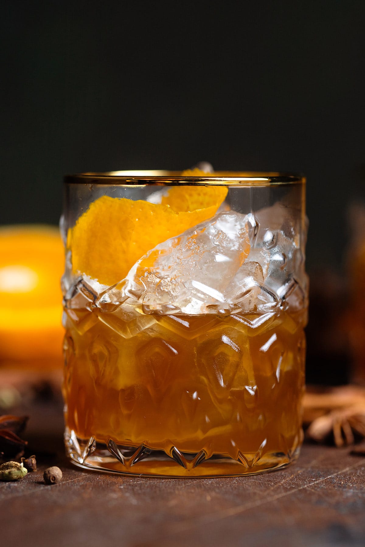 Old fashioned cocktail in a gold-rimmed glass with ice, orange zest, and star anise.