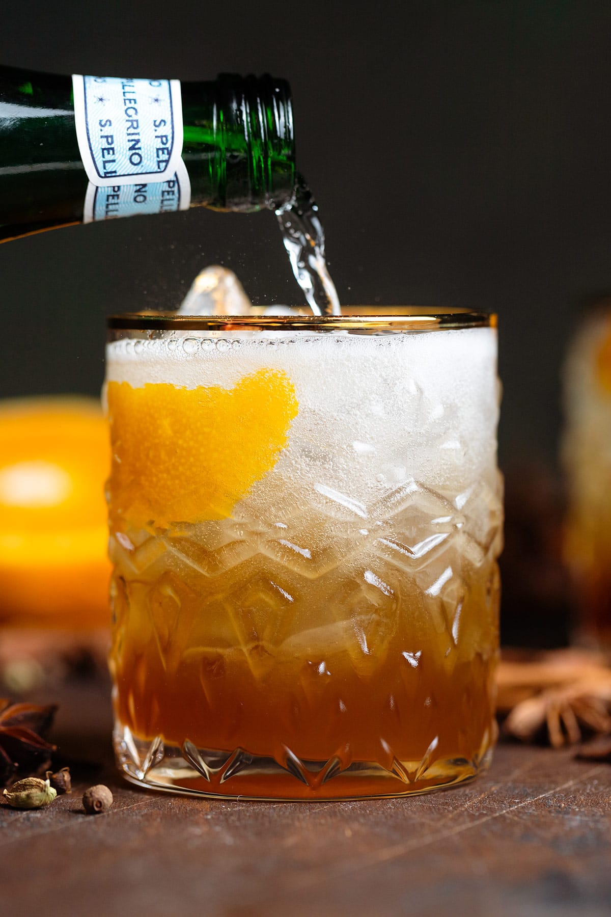 Old fashioned cocktail in a gold-rimmed glass with ice, orange zest, and star anise being topped with sparkling water.