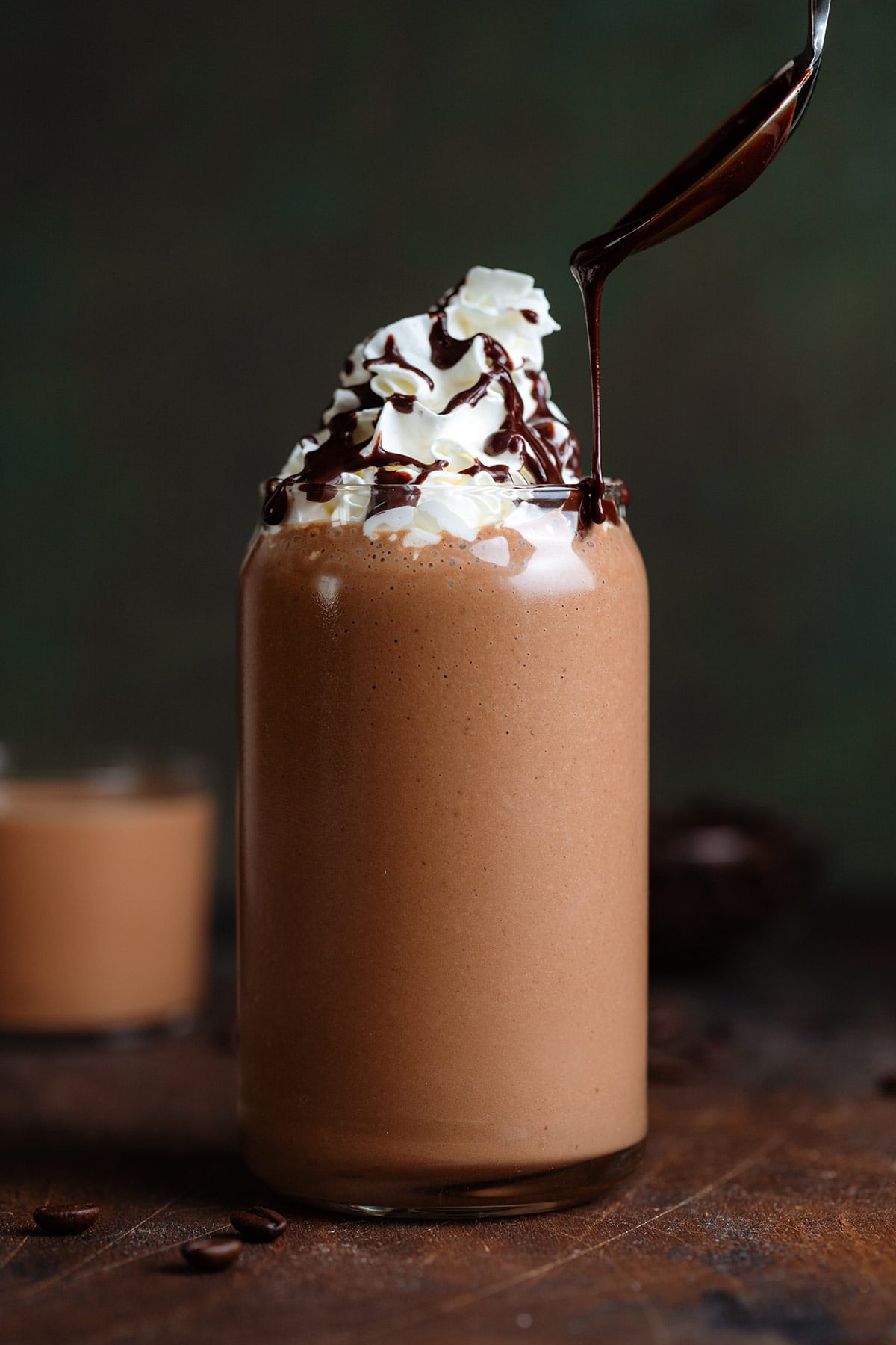 A dark brown mocha smoothie in a tall glass garnished with whipped cream being drizzled with chocolate sauce.