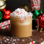 White chocolate mocha in a glass mug with whipped cream and crushed candy canes on top and more whole candy canes and Christmas ornaments in the background.