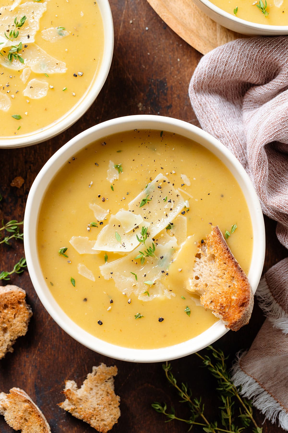 Light yellow potato soup in a white bowl garnished with parmesan slices, black pepper, fresh thyme, and toasted bread dipped into the soup.