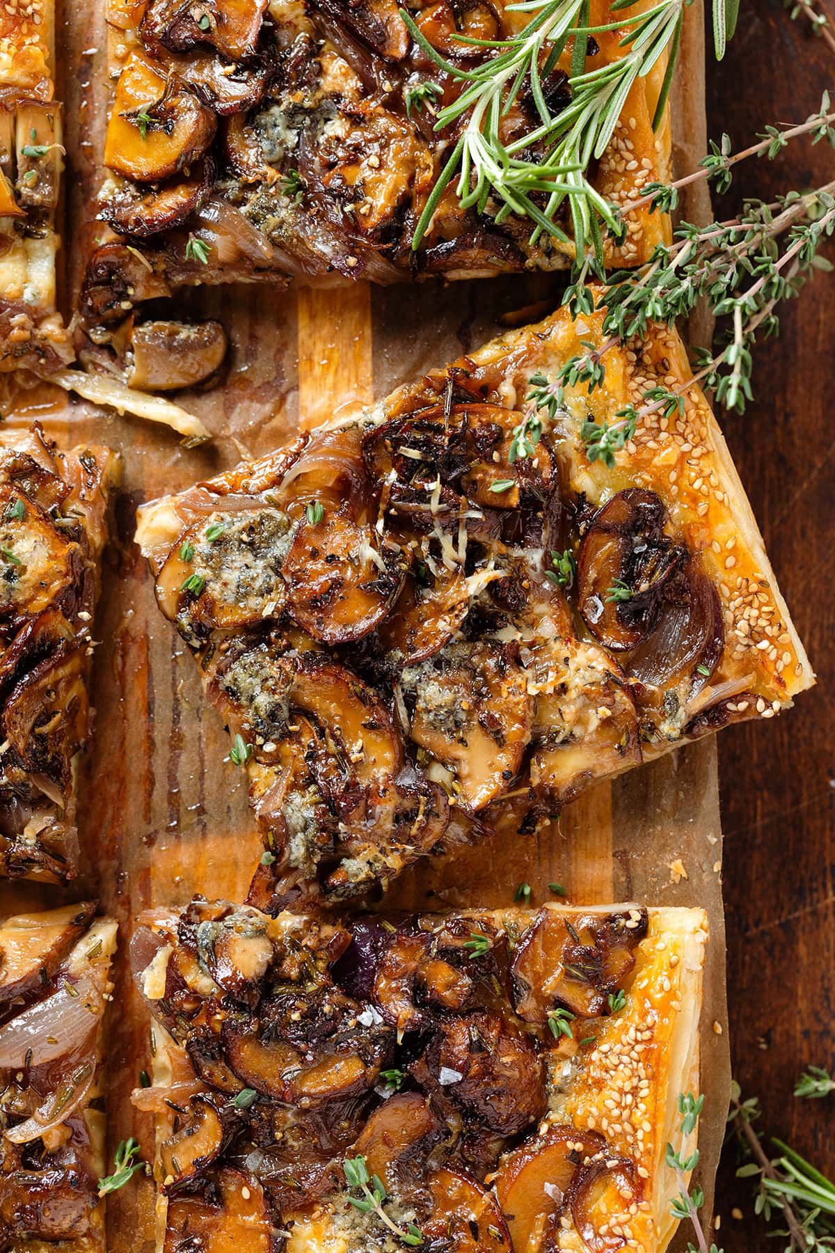 Baked puff pastry mushroom tart sliced into squares on a wooden cutting board topped with fresh thyme and rosemary.