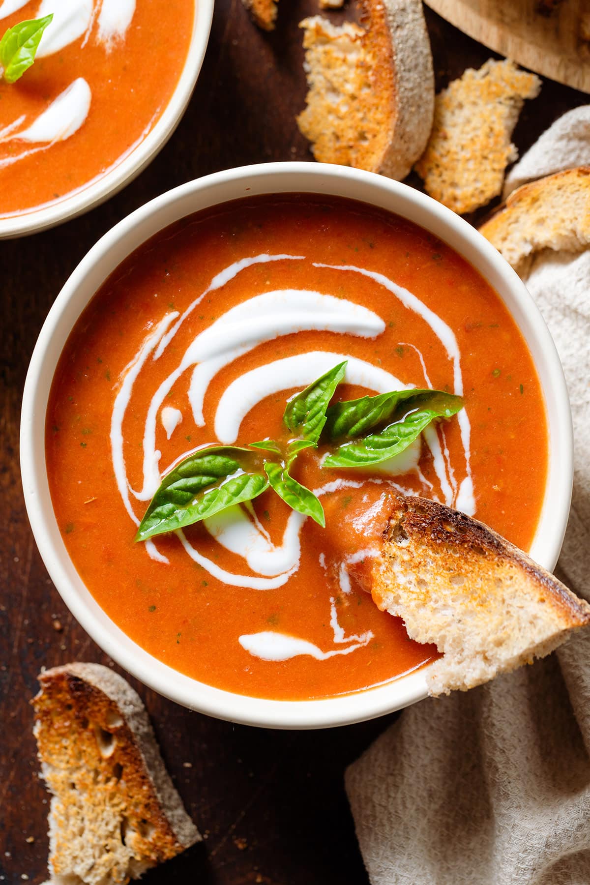 Tomato soup with a drizzle of yogurt in a white bowl garnished with fresh basil and a piece of toasted bread dipped into it.