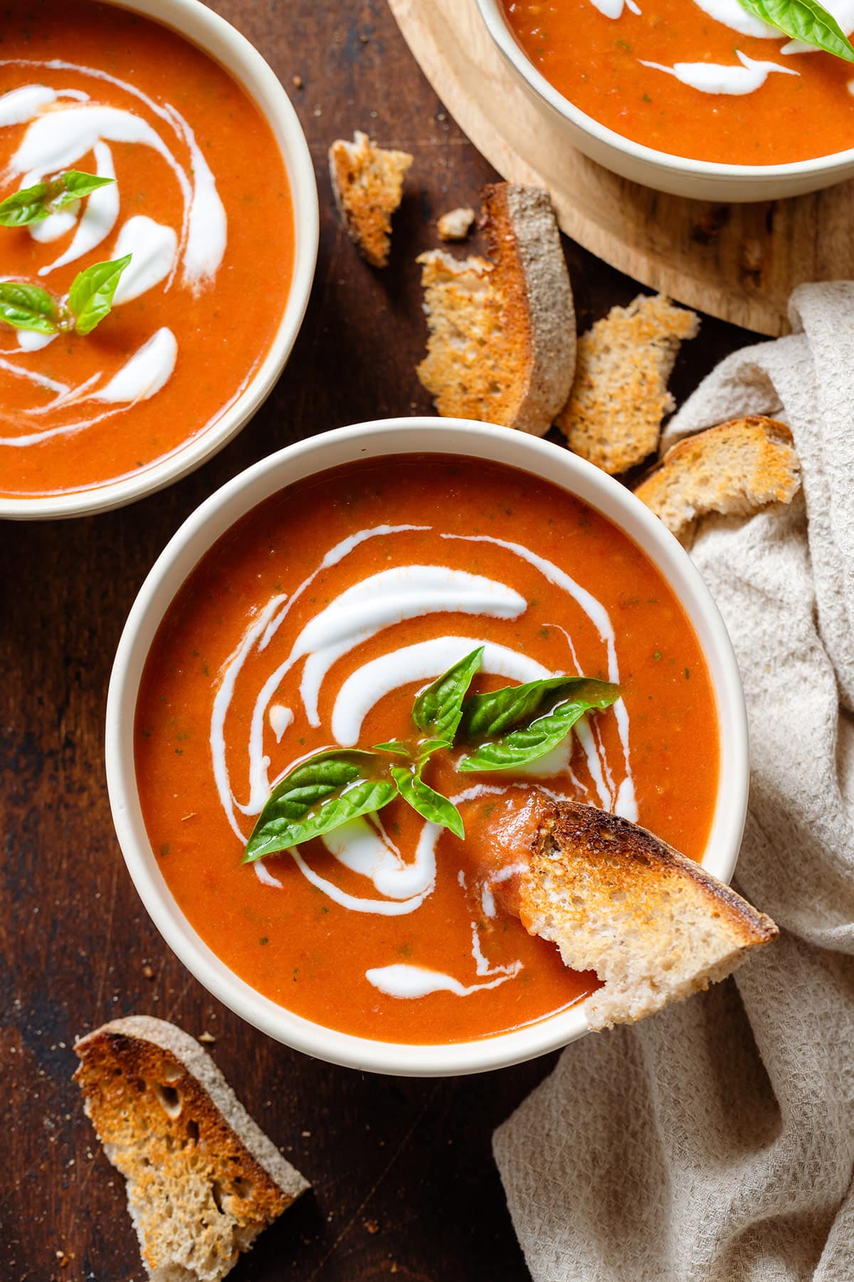Tomato soup with a drizzle of yogurt in a white bowl garnished with fresh basil and a piece of toasted bread dipped into it.