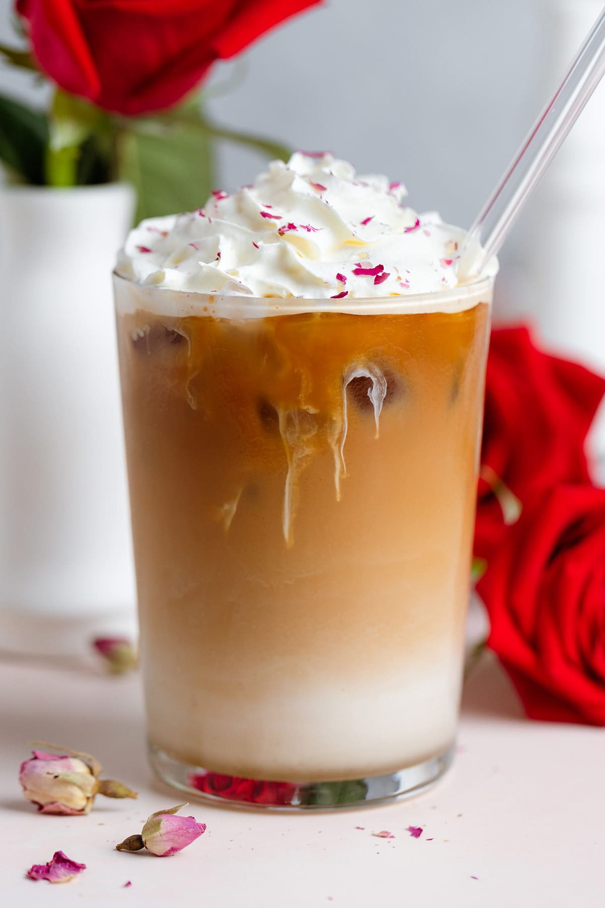 Iced latte with whipped cream and crushed dried rose petals sprinkled on top with a glass straw with red roses around the glass.