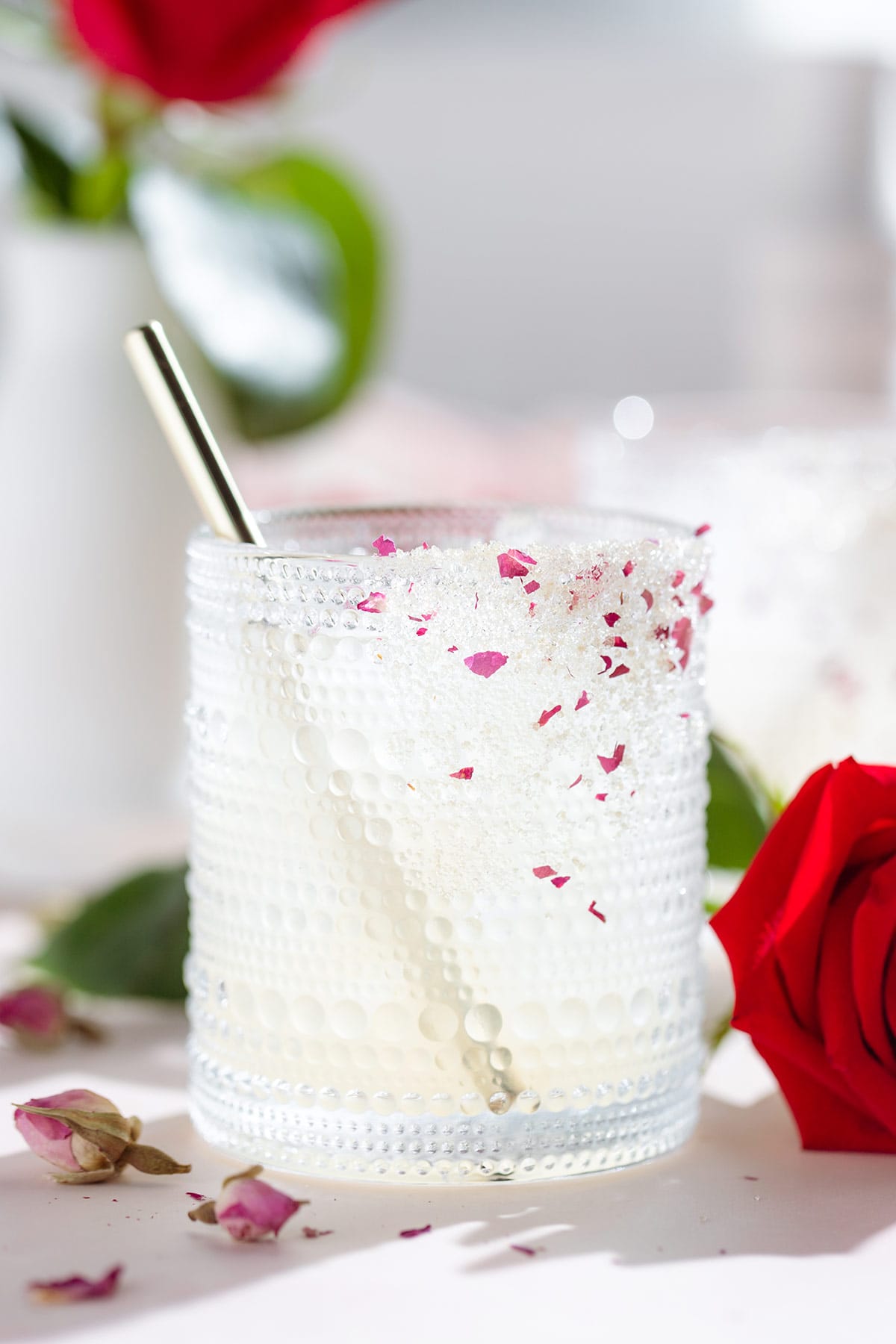 A margarita cocktail in a short glass garnished with a sugar rim with crushed dried rose petals and a gold straw with red roses around the glass.