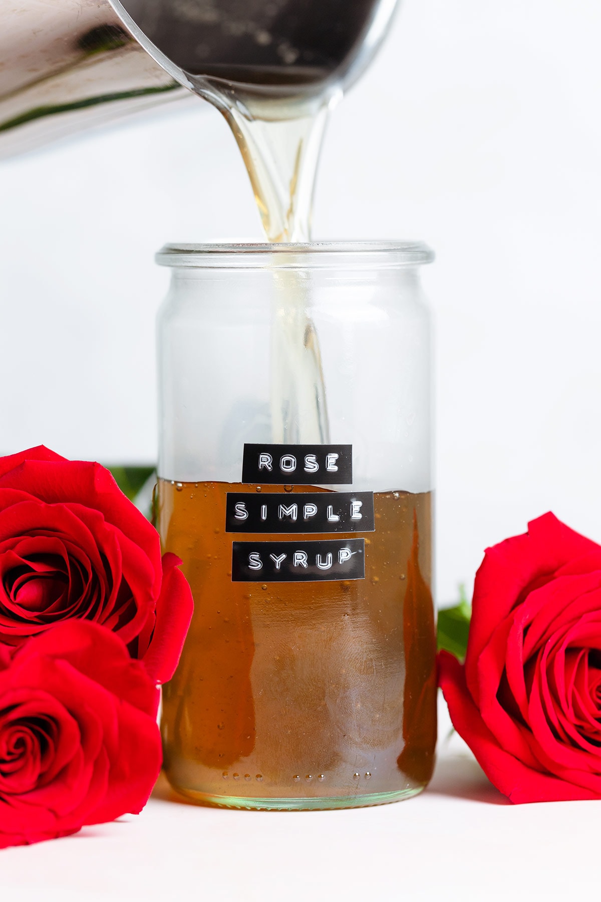 Syrup being poured into a glass jar that has a jar that says rose simple syrup on it with red roses around the jar.