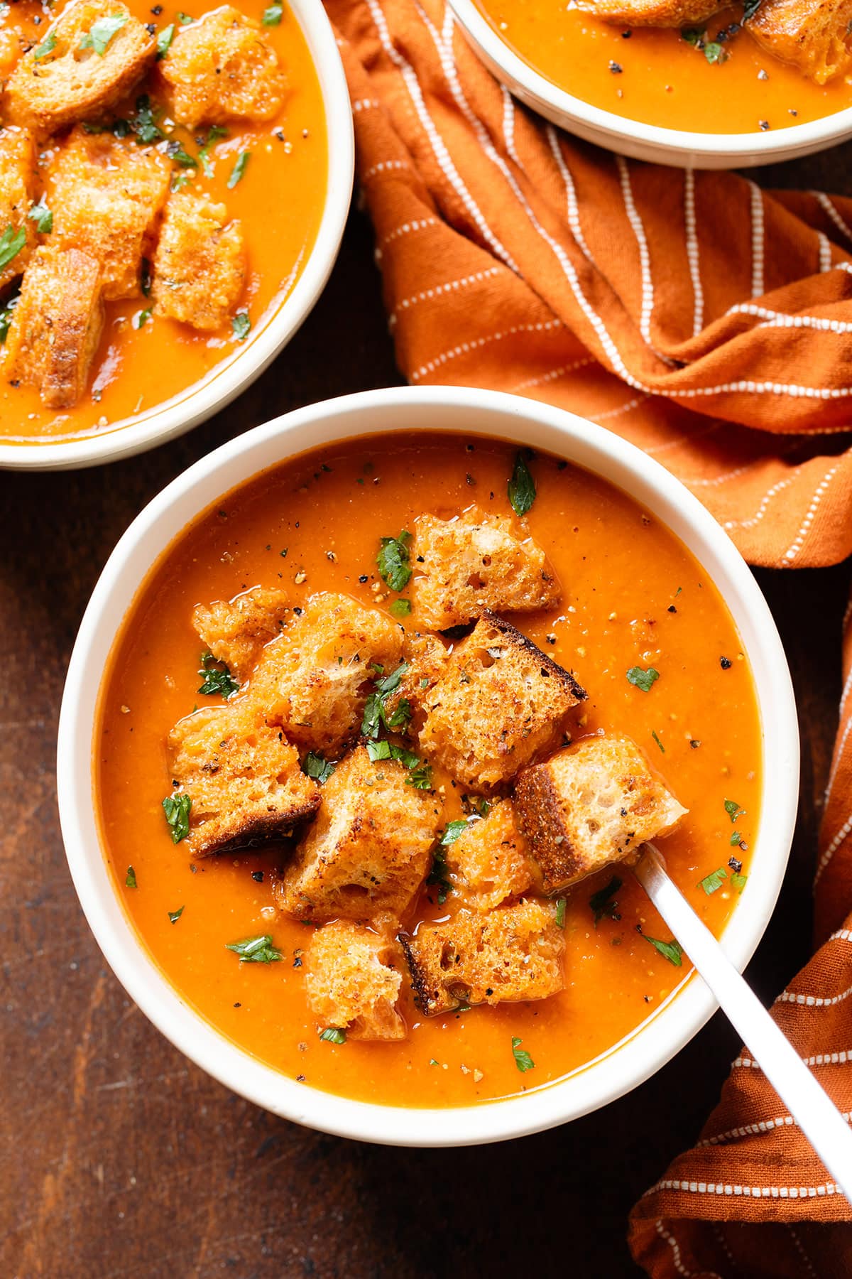 Tomato soup in white bowls with a spoon topped with croutons, fresh parsley, and black pepper.