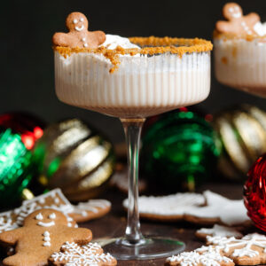 A creamy beige gingerbread martini in a coupe glass garnished with crushed cookies around the rim and whipped cream and a small gingerbread man on top.