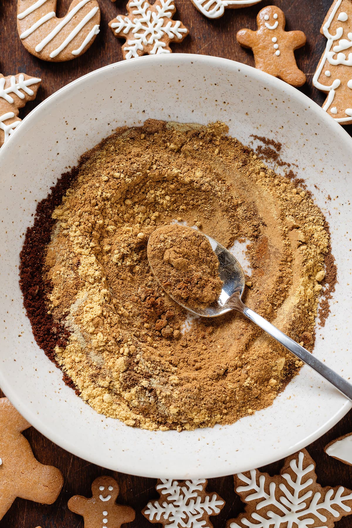 Various spices in a beige low bowl being stirred together with a spoon to make gingerbread spice.
