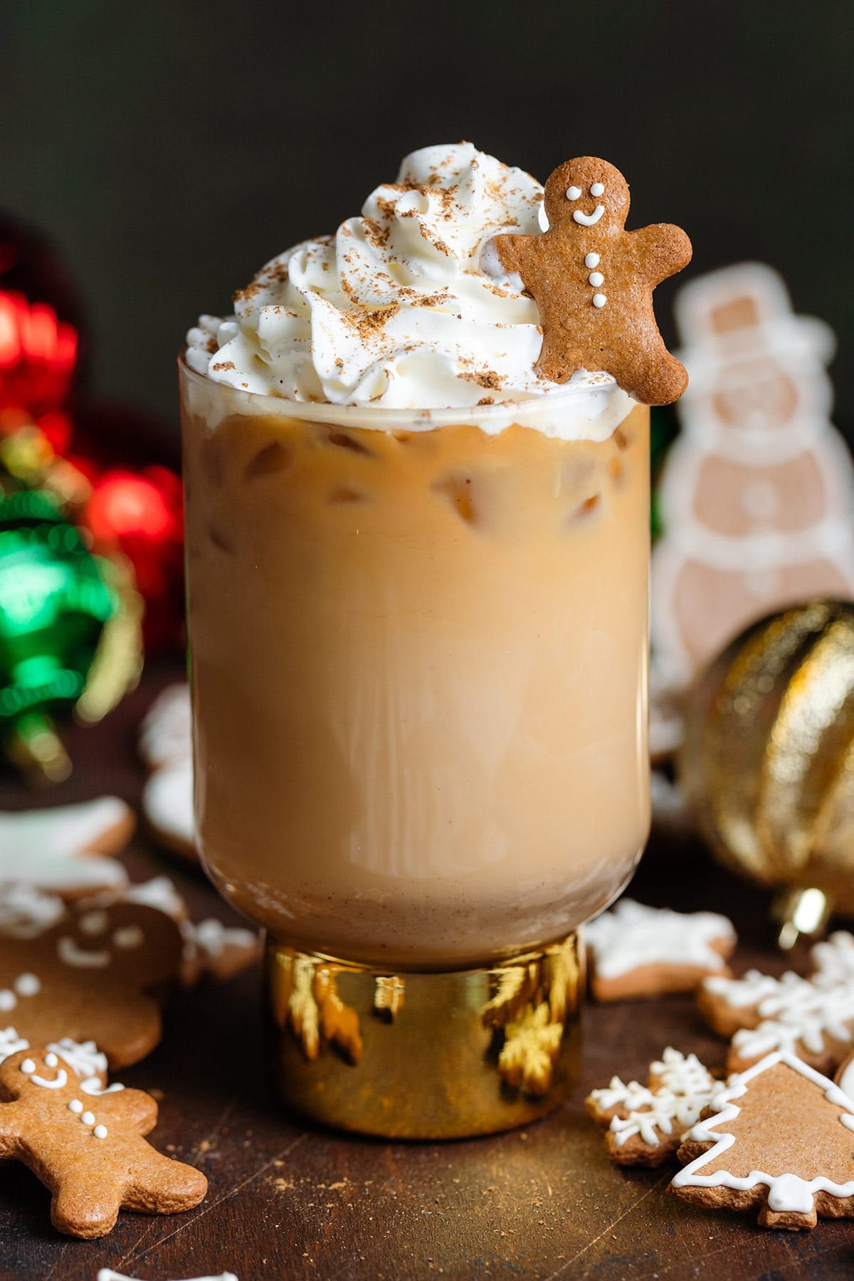 An iced gingerbread latte in a tall glass with a gold bottom topped with whipped cream, gingerbread spice, and a small gingerbread man cookie.