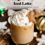 An iced gingerbread latte in a tall glass with a gold bottom topped with whipped cream, gingerbread spice, and a small gingerbread cookie.
