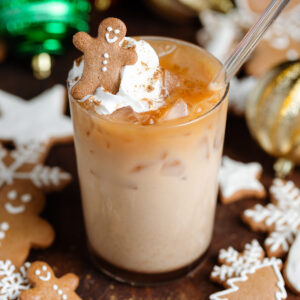 Iced chai latte in a tall glass topped with a dollop of whipped cream, gingerbread spice, and a small gingerbread man with a glass straw.