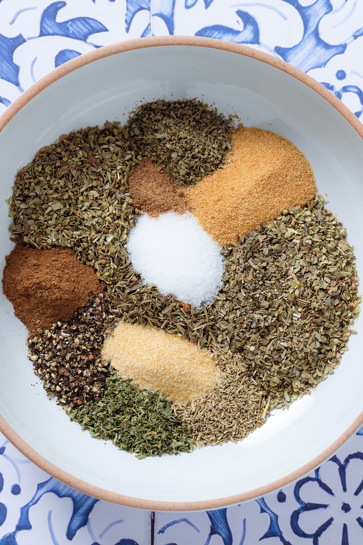 Dried herbs and spices neatly laid out on a white bowl on a blue background.