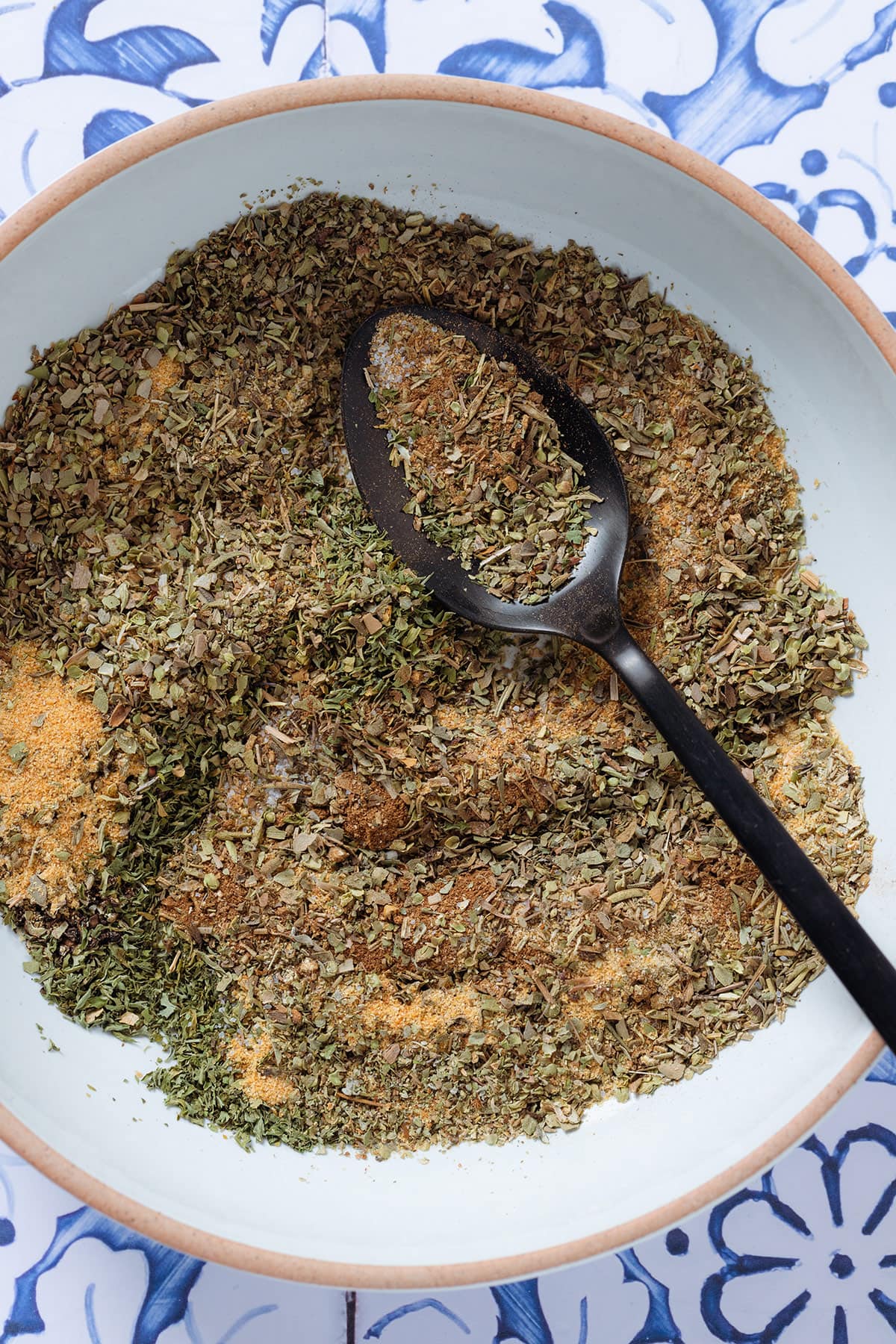 Dried herbs and spices in a white bow partially mixed with a black spoon.