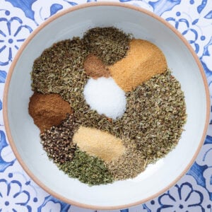Dried herbs and spices neatly laid out on a white bowl on a blue background.