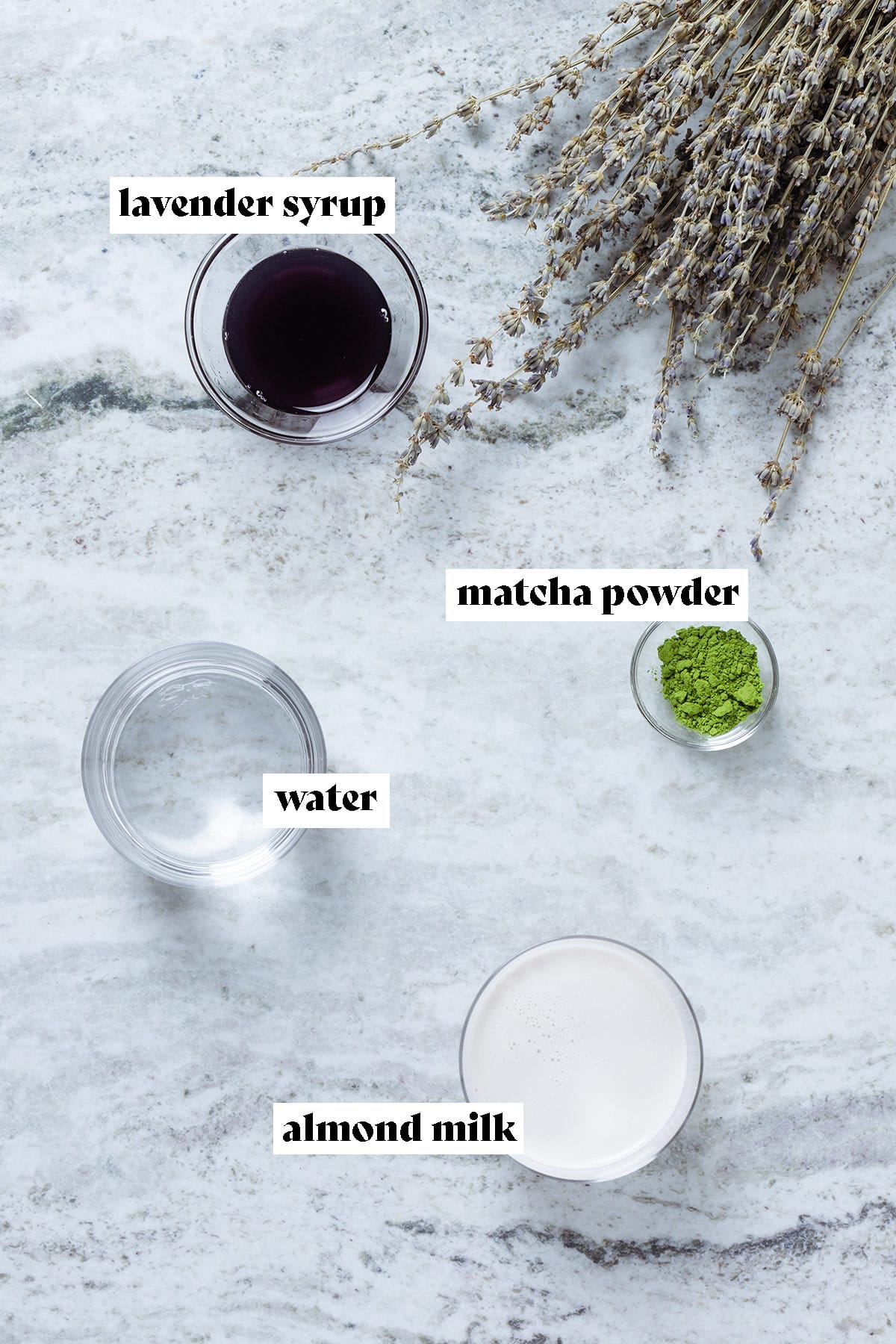 Measured out matcha powder, milk, water, and lavender syrup laid out in bowls on a grey stone background.
