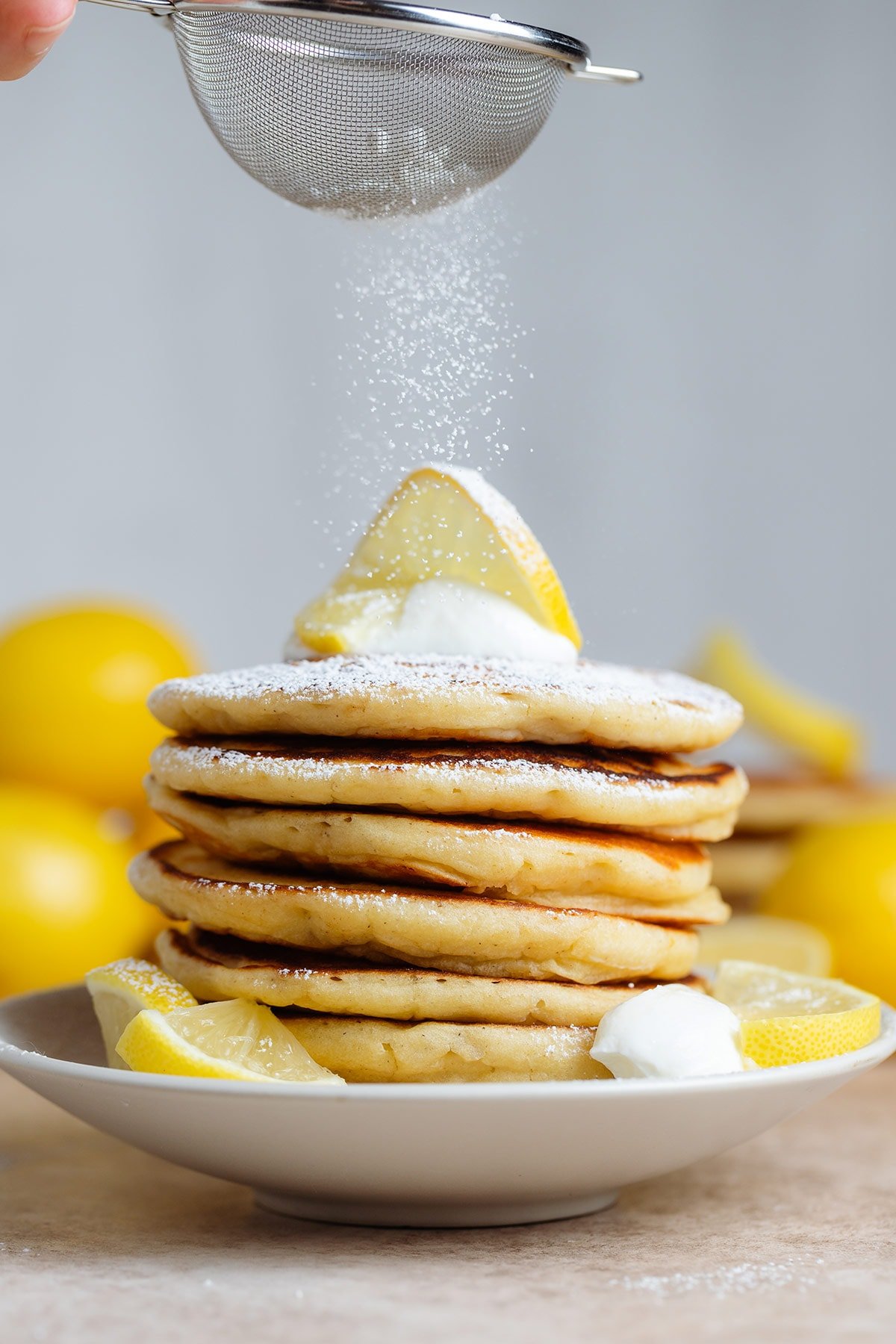 A stack of pancakes with lemon slice on top and a dollop of yogurt being dusted with powdered sugar.