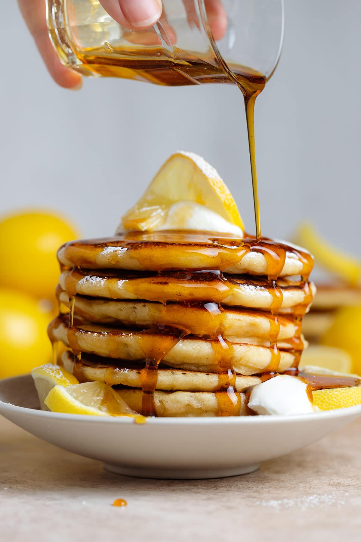 A stack of pancakes with yogurt and lemon slices being drizzled with maple syrup.
