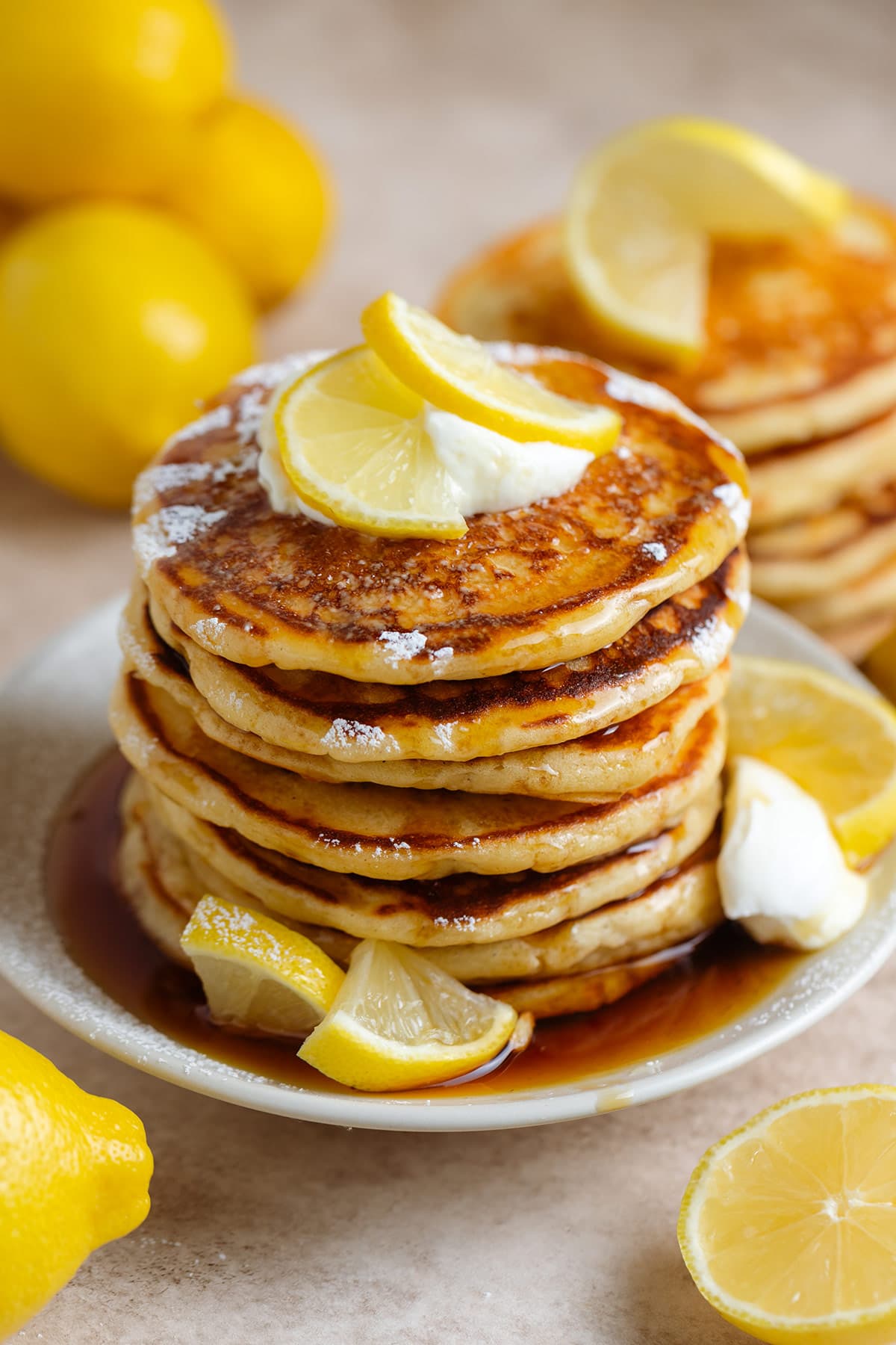 A stack of pancakes garnished with lemon slices, yogurt, and maple syrup on top and on the sides.