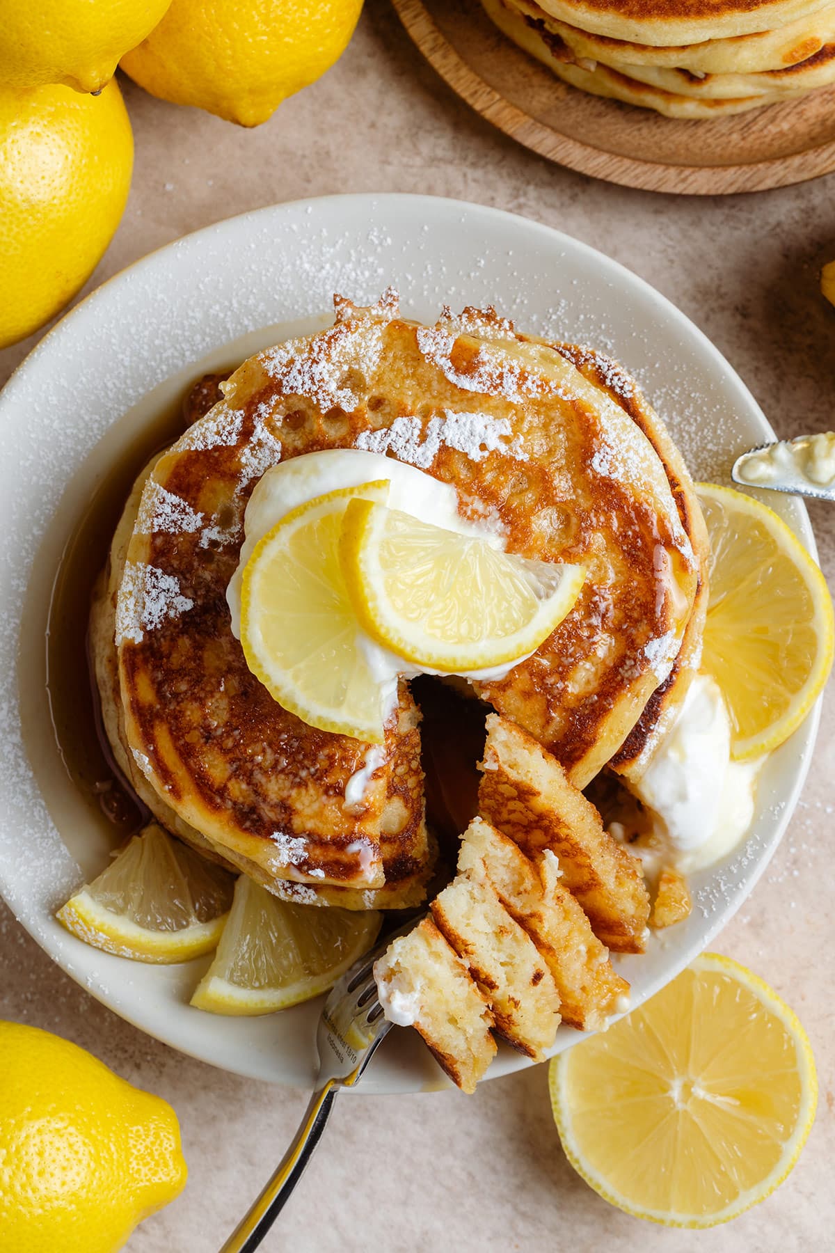 A stack of pancakes garnished with lemon slices, yogurt, and maple syrup with a piece cut out and resting on the side on a fork.