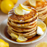 A stack of pancakes garnished with lemon slices, yogurt, and maple syrup on top and on the sides.