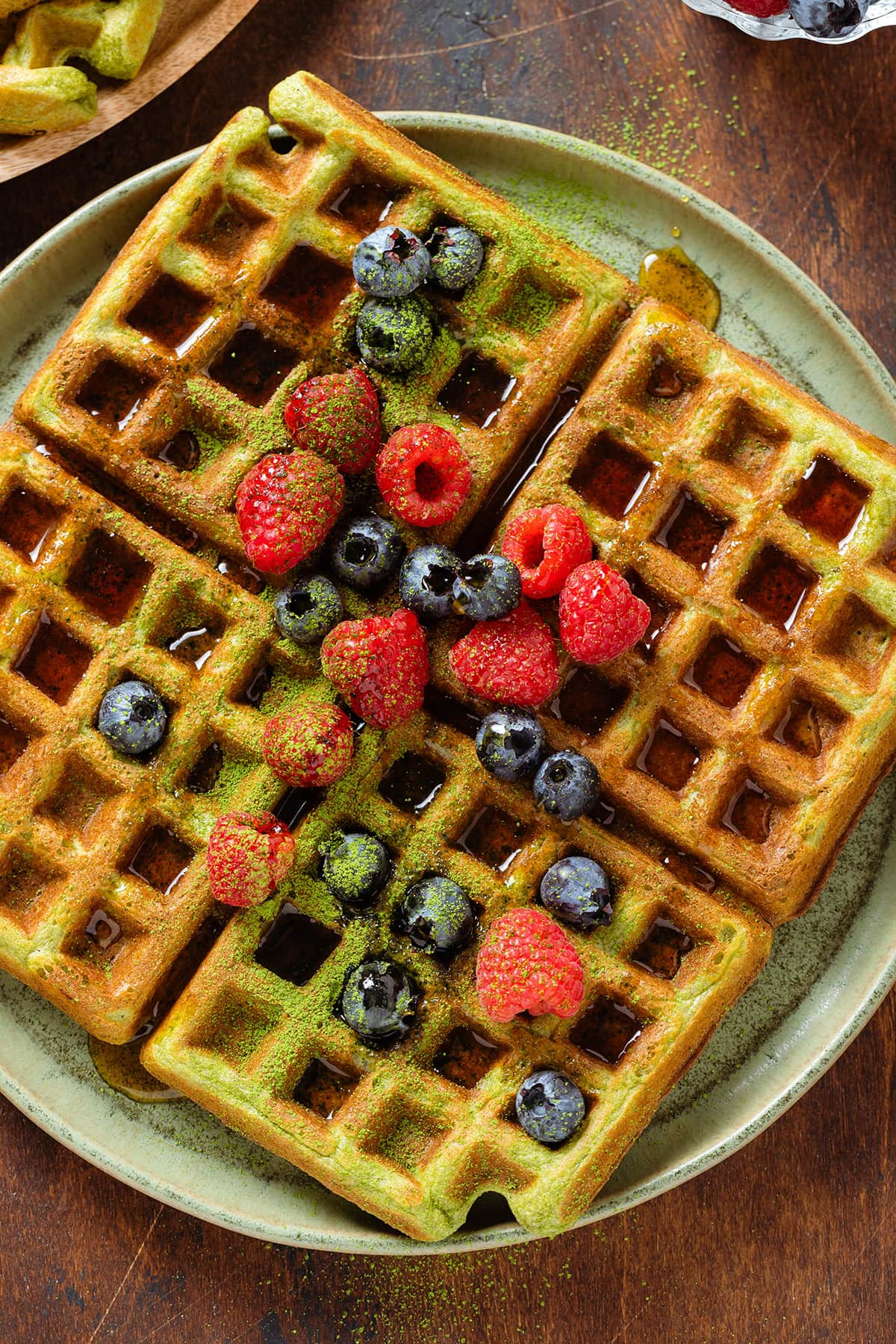 Greek waffles on a green plate garnished with fresh berries, maple syrup, and dusted with matcha powder.