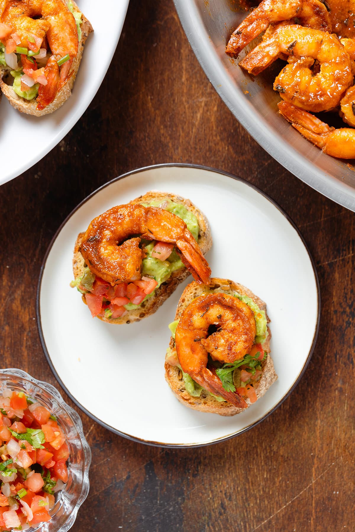 Toasted baguette slices topped with guacamole, fresh salsa, and cajun shrimp on a small plate.
