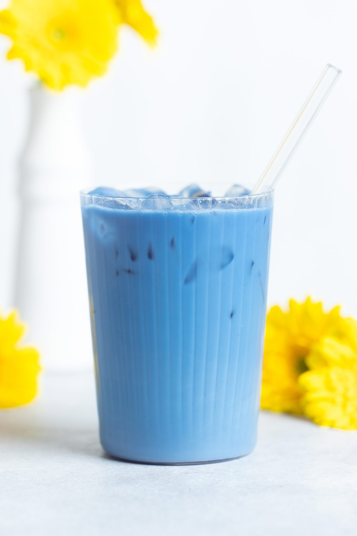 Bright blue iced latte in a tall ribbed glass with a glass straw and yellow flowers around the glass.
