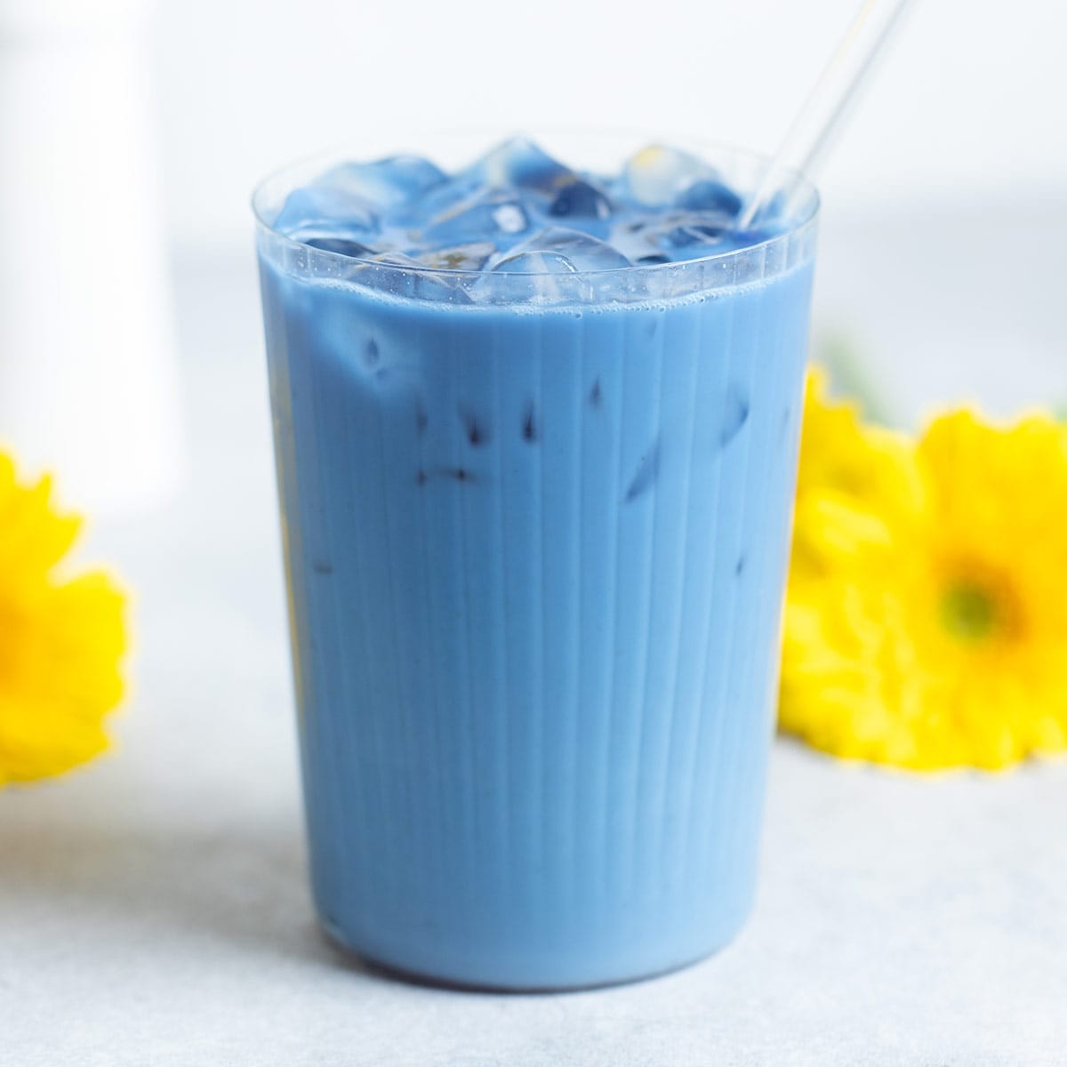Bright blue iced latte in a tall ribbed glass with a glass straw and with yellow flowers around the glass.