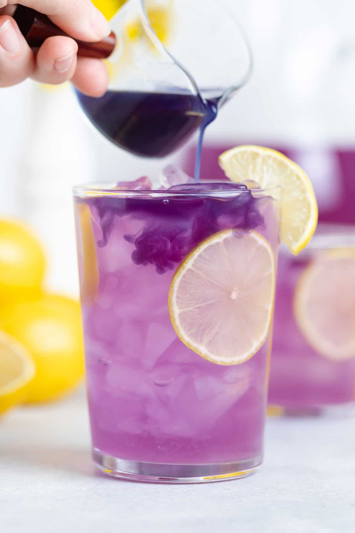 Blue matcha being slowly drizzled into a tall glass with purple lemonade garnished with lemon slices.