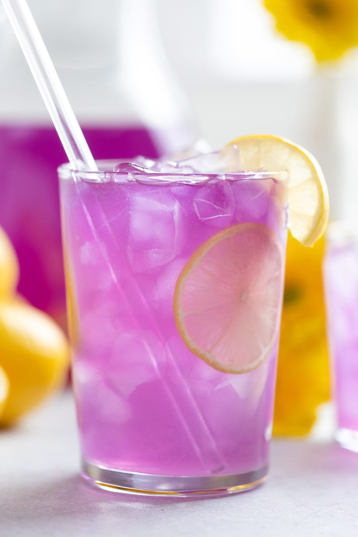 Bright purple lemonade in a tall glass with ice garnished with lemon slices and a glass straw.