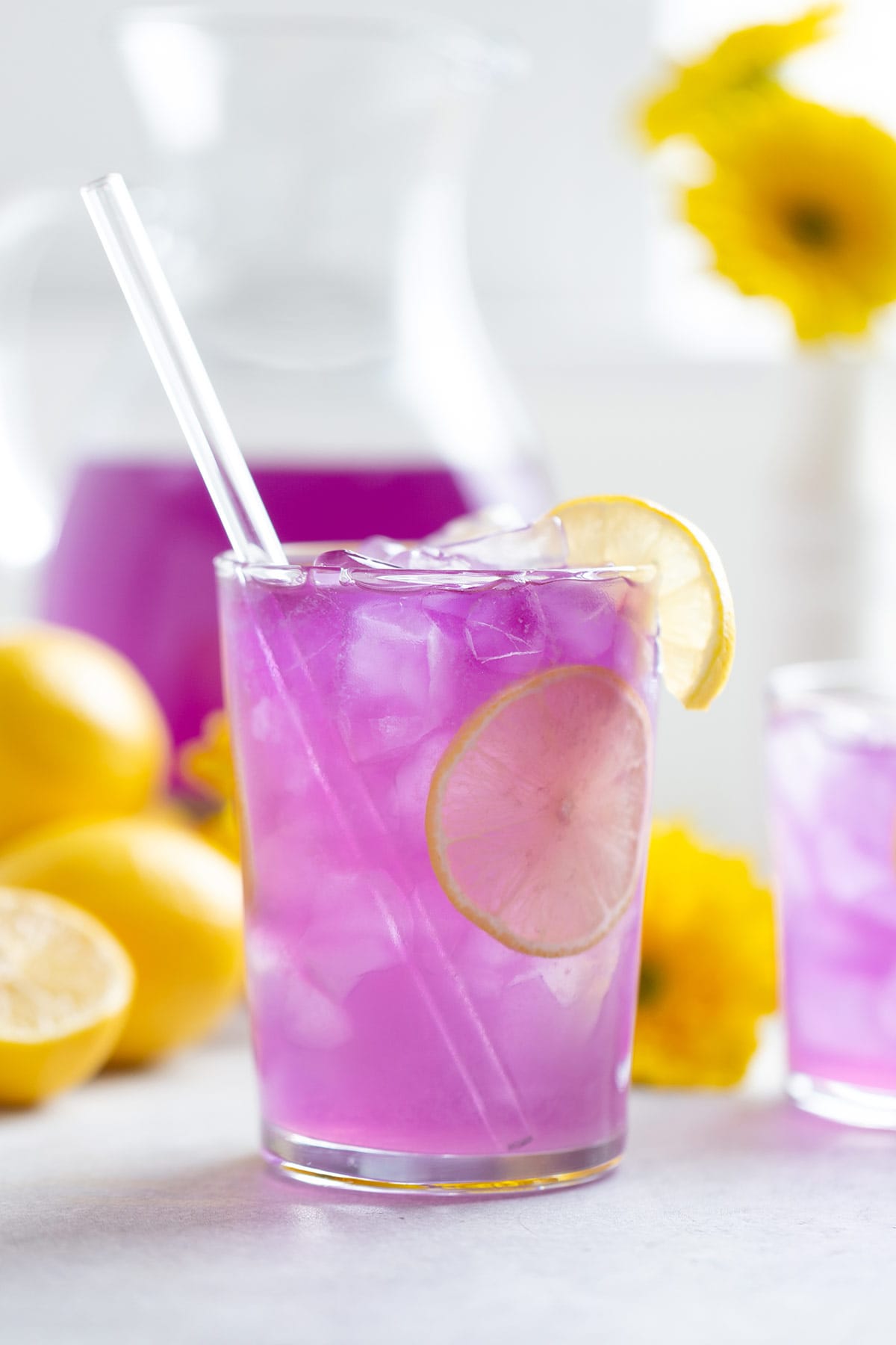 Bright purple lemonade in a tall glass with ice garnished with lemon slices and a glass straw.