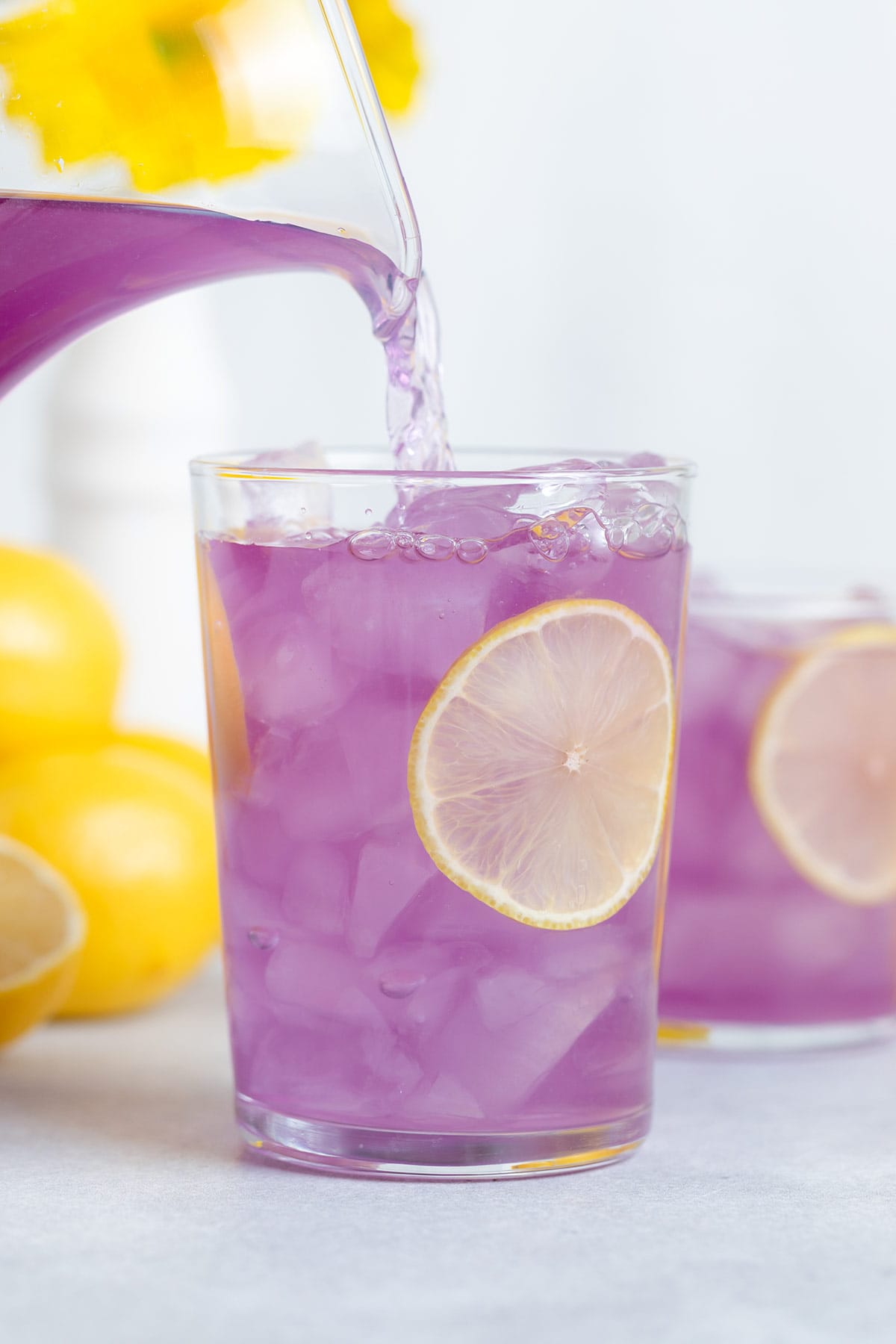 Purple lemonade being poured over ice into a tall glass garnished with lemon slices.