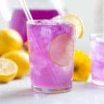 Bright purple lemonade in a tall glass with ice garnished with lemon slices and a glass straw.