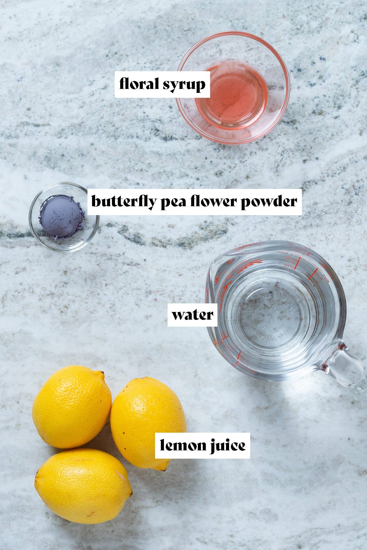 Lemons, blue powder, water, and pink syrup measured in bowls all laid out on a grey background with text overlay.