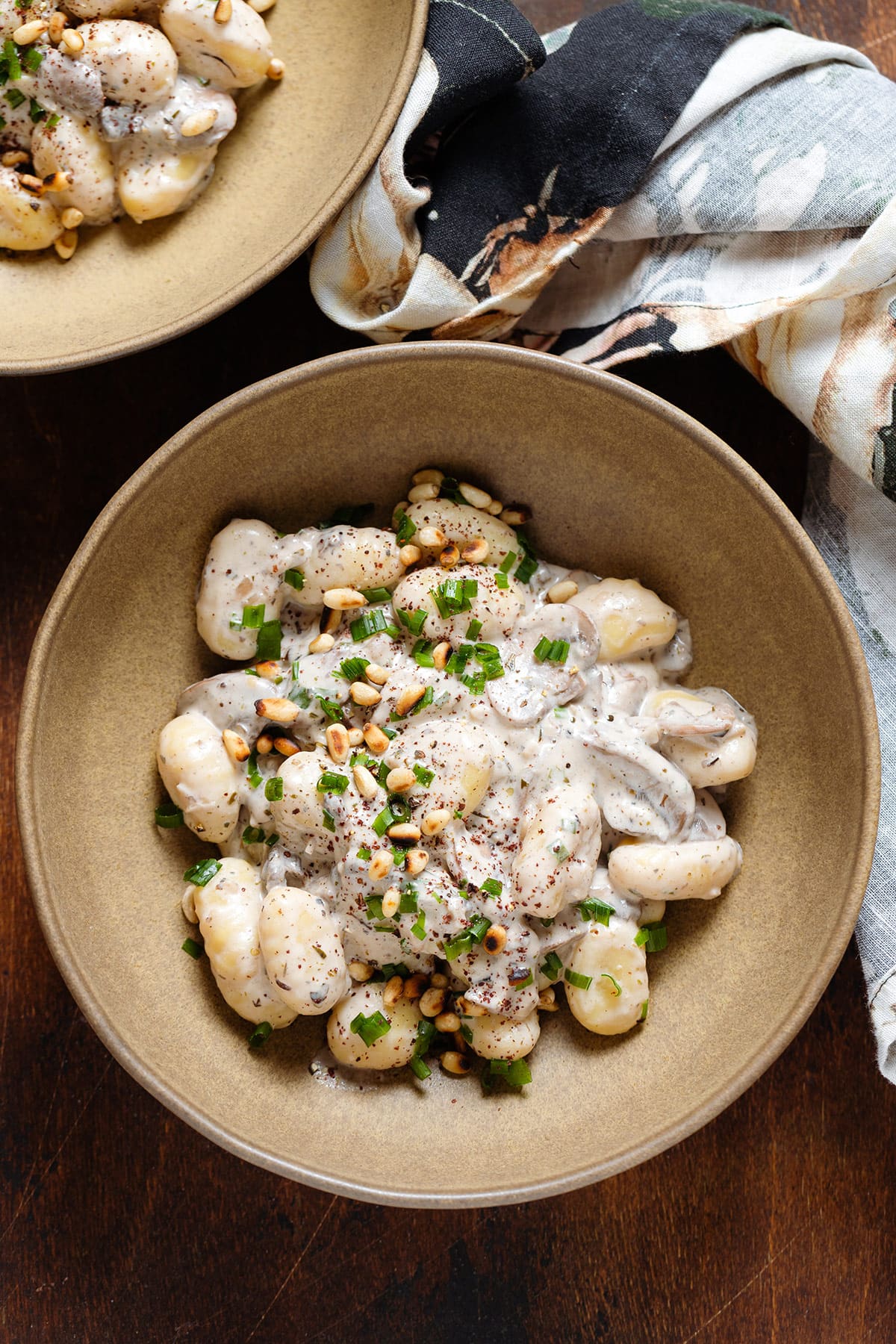 Creamy gnocchi with mushroom in a low brown bowl topped with toasted pine nuts, chives, and sumac.
