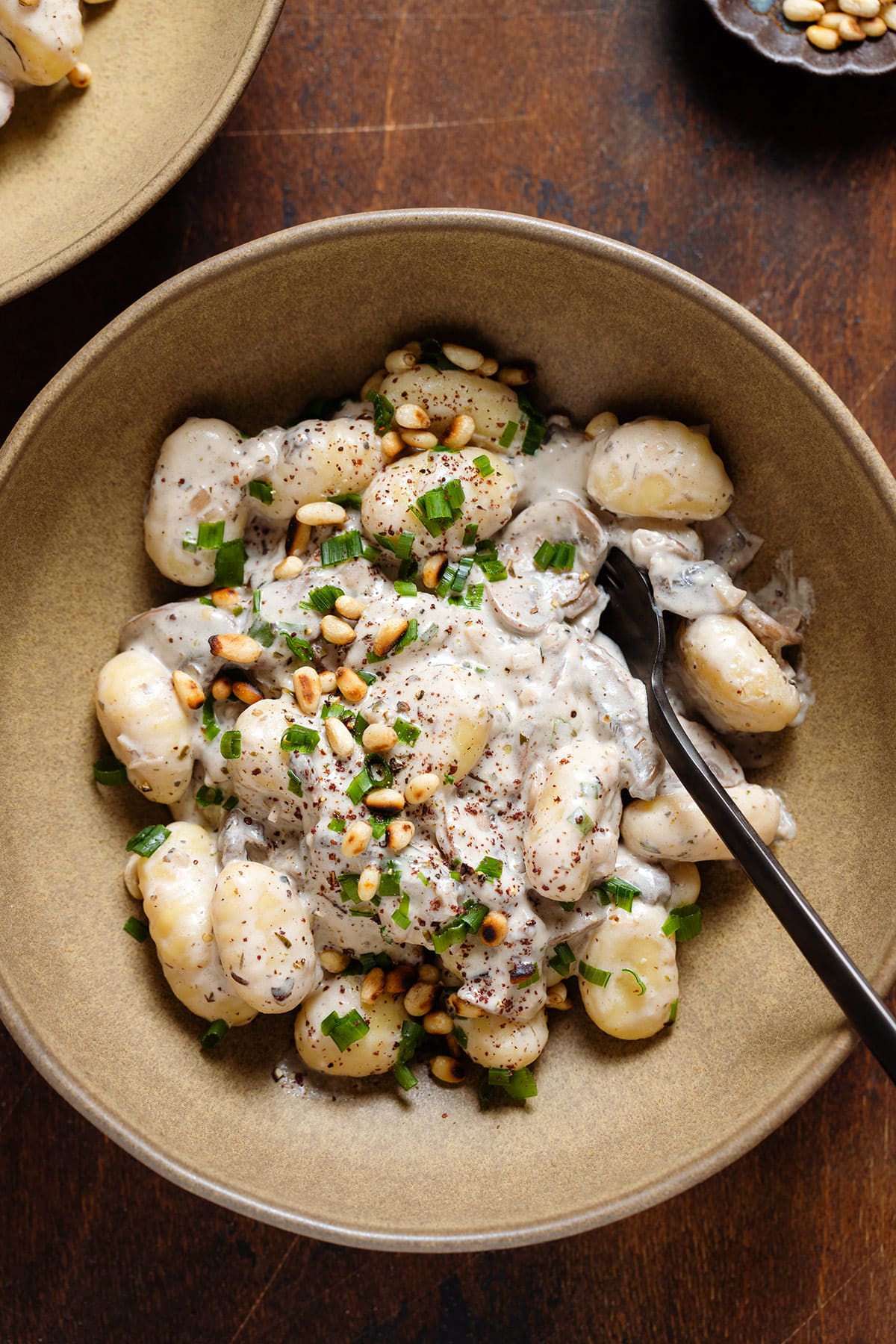 Creamy gnocchi with mushroom in a low brown bowl topped with toasted pine nuts, chives, and sumac with a black fork on the right side of the bowl.