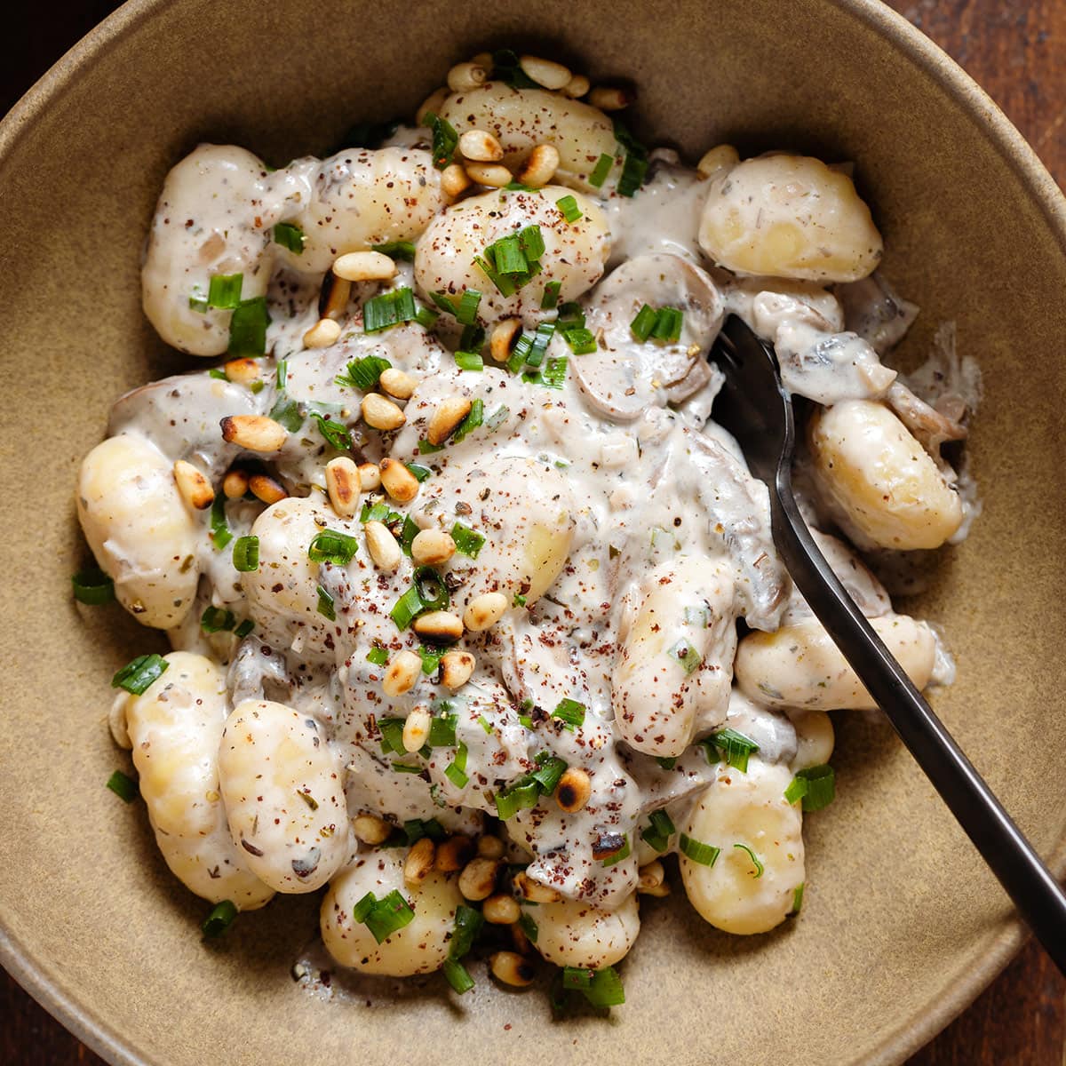 Creamy gnocchi with mushroom in a low brown bowl topped with toasted pine nuts, chives, and sumac with a black fork on the right side of the bowl.
