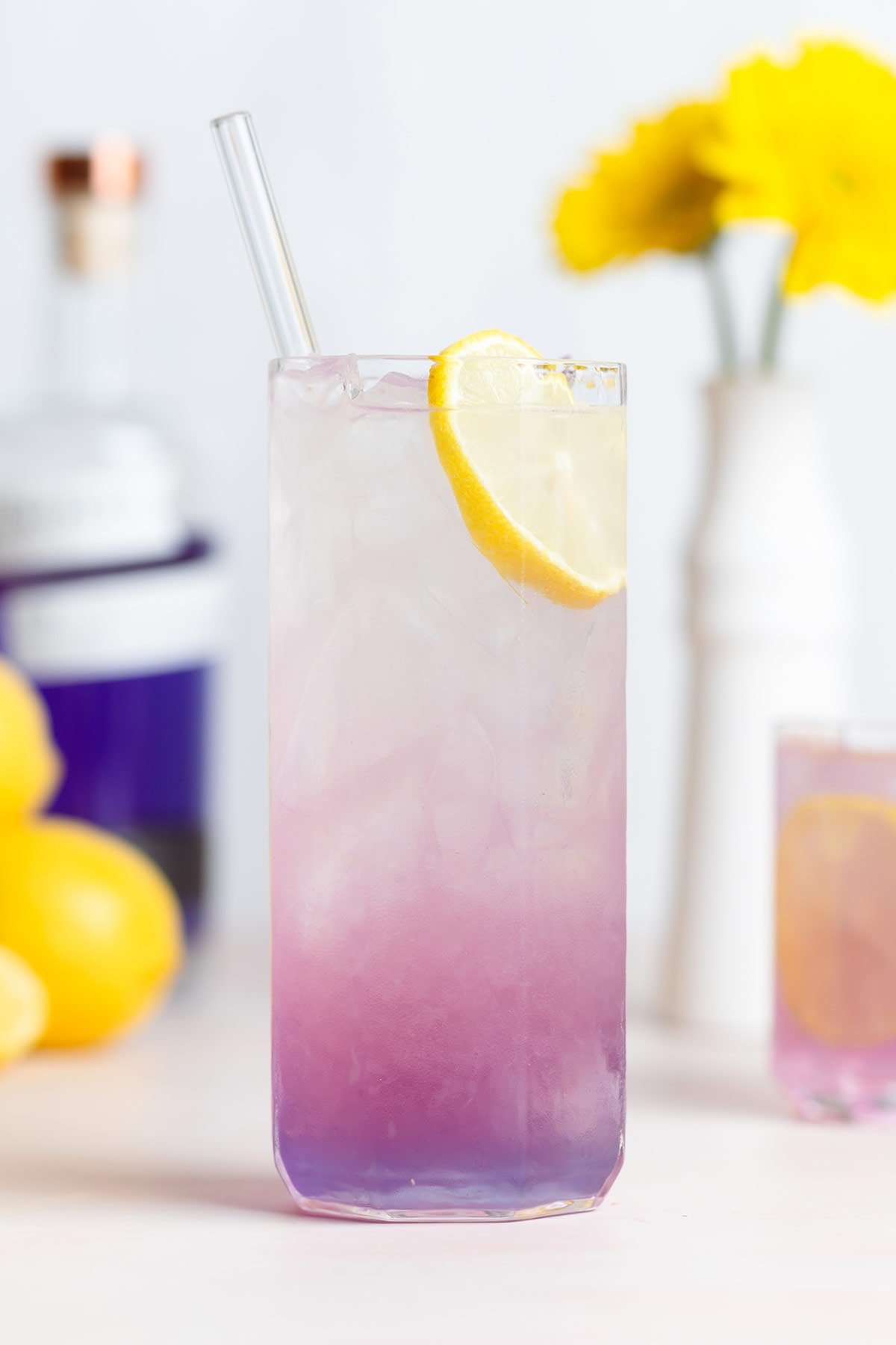 A light pink purple cocktail in a tall glass with an ombre effect garnished with a slice of lemon and a glass straw.